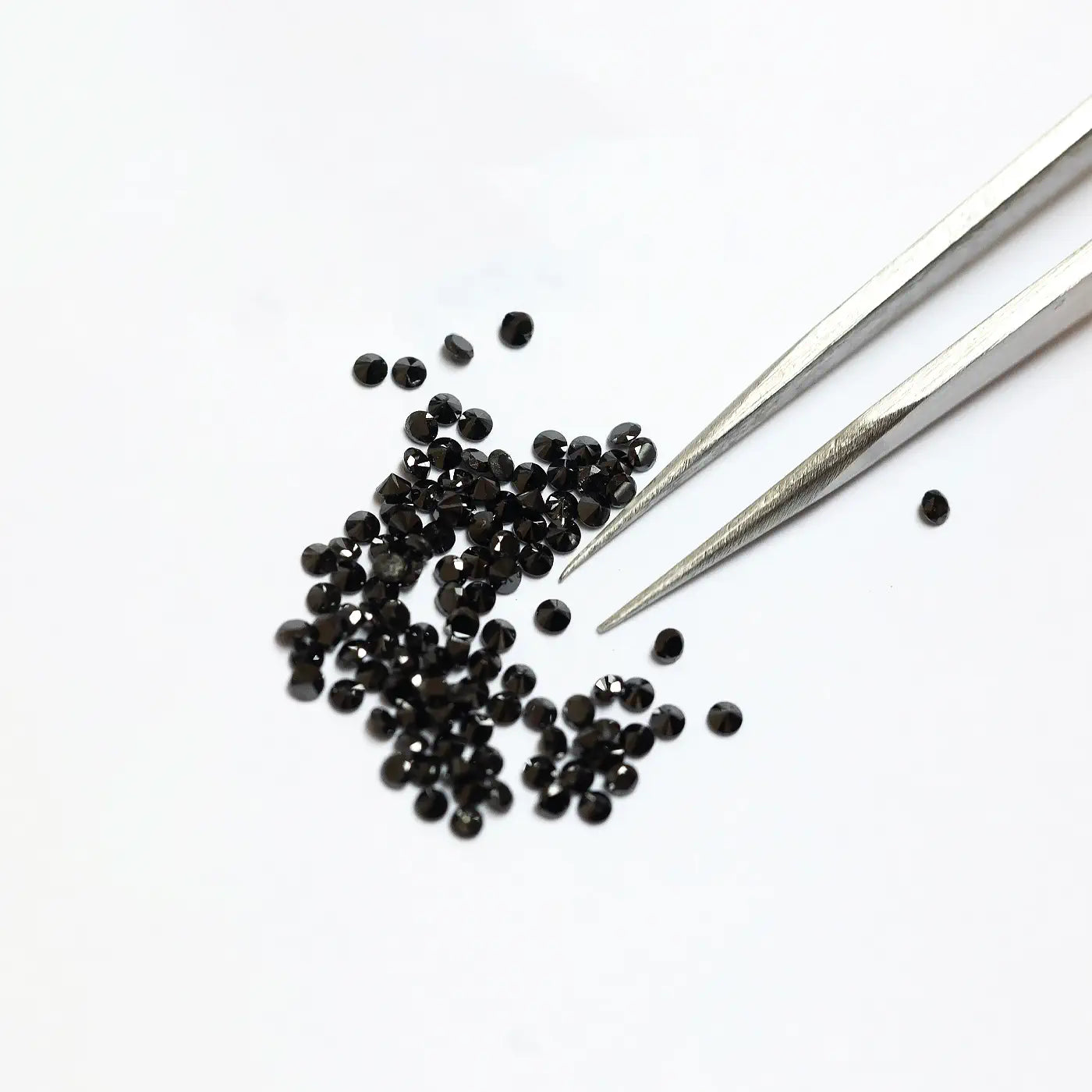 Gros plan sur des pinces à côté d’un tas éparpillé de petits diamants noirs ronds sur une surface blanche.