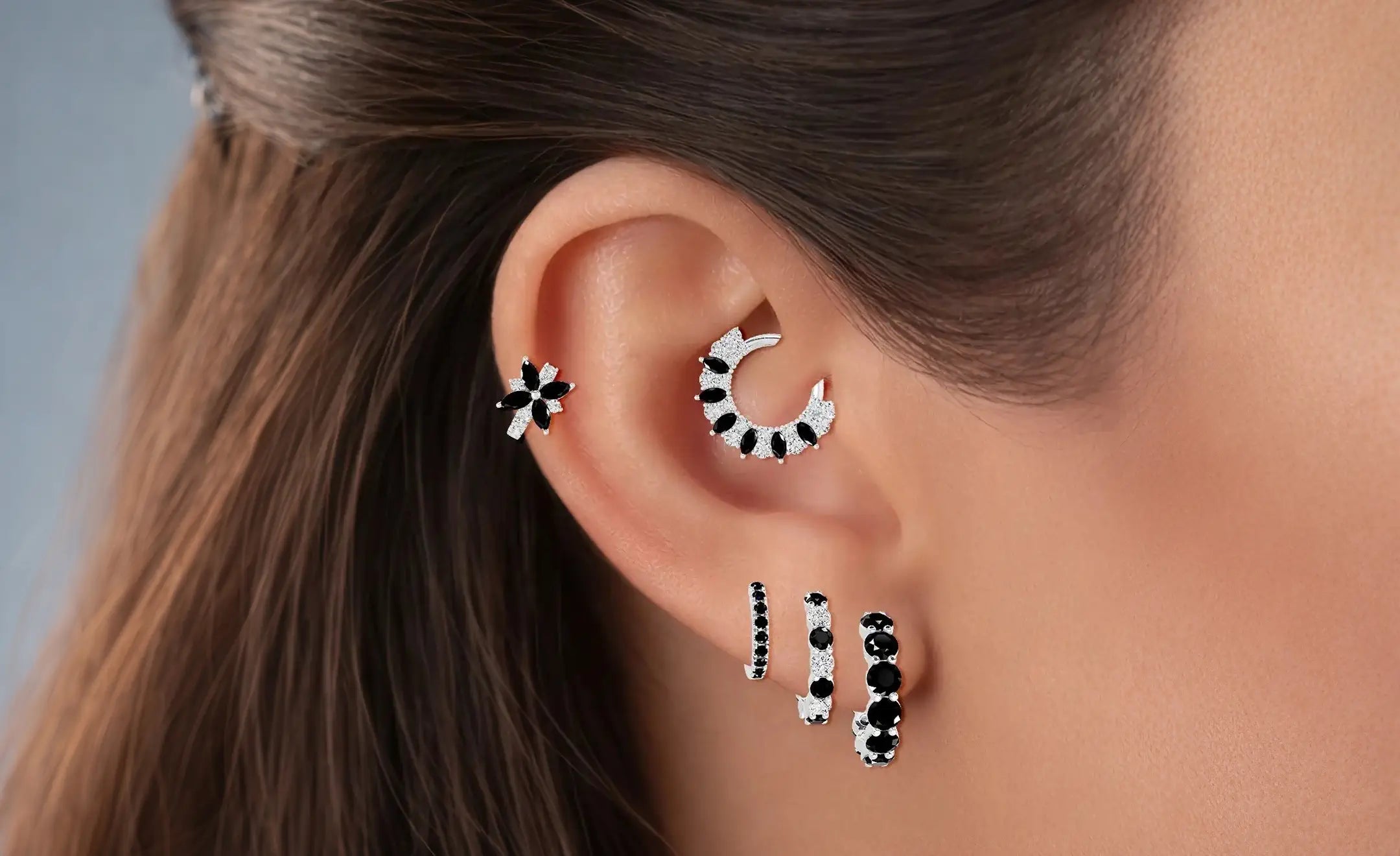 Ear stack of a women wearing diamond and black diamond in 18k white gold. A graduated hoop piercing on her lobe, flower black diamond hoop on the helix and a combination of black diamond and diamond daith piercing 
