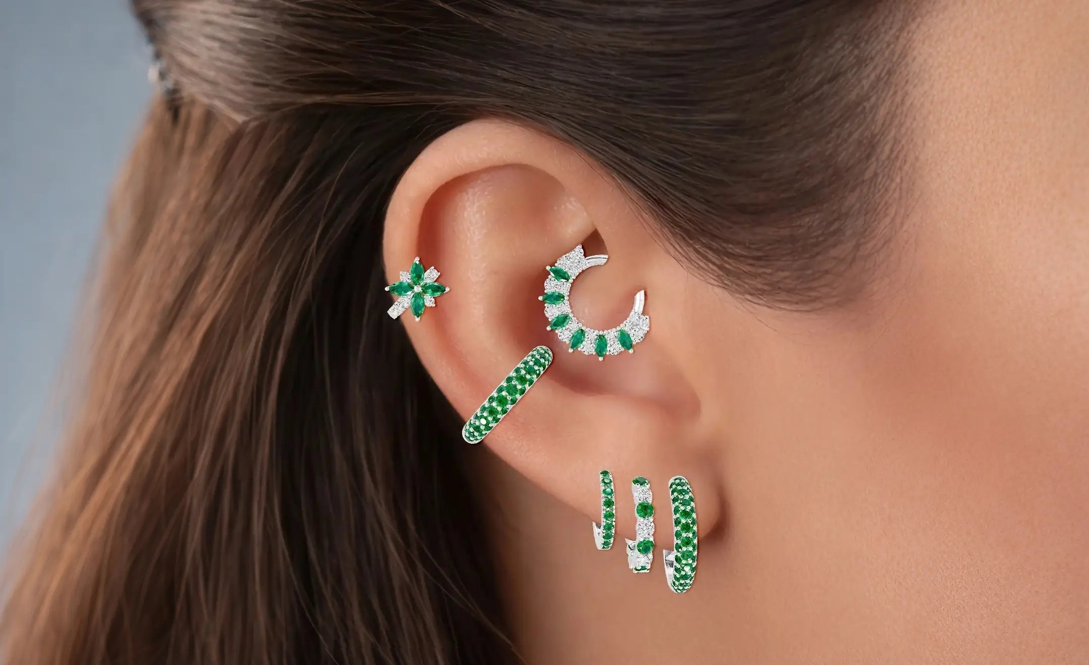 Emerald and diamond ear piercing in an ear stack. Three descending hoop earrings on the lobe, a pave emerald piercing, a flower diamond and emerald hoop on the middle helix, and last is a combination of diamond an emerald helix 