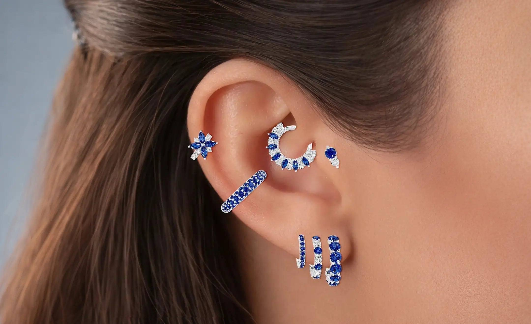 Sapphires and diamonds ear stack in a woman's ear. A sapphire and diamond hoop in descending order. A pave hoop piercing on the lower helix and a sapphire flower hoop piercing. One combination of sapphire and diamond on the daith and tragus