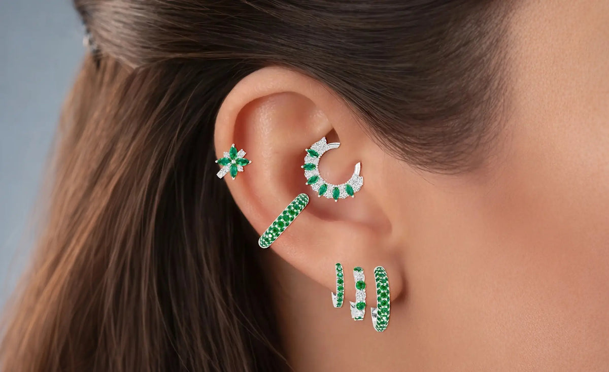 Composition d'oreille avec piercings émeraude et diamant. Trois créoles décroissantes sur le lobe, un piercing émeraude pavé, une créole hélix fleur diamant et émeraude au milieu, et un piercing hélix combinant diamant et émeraude.