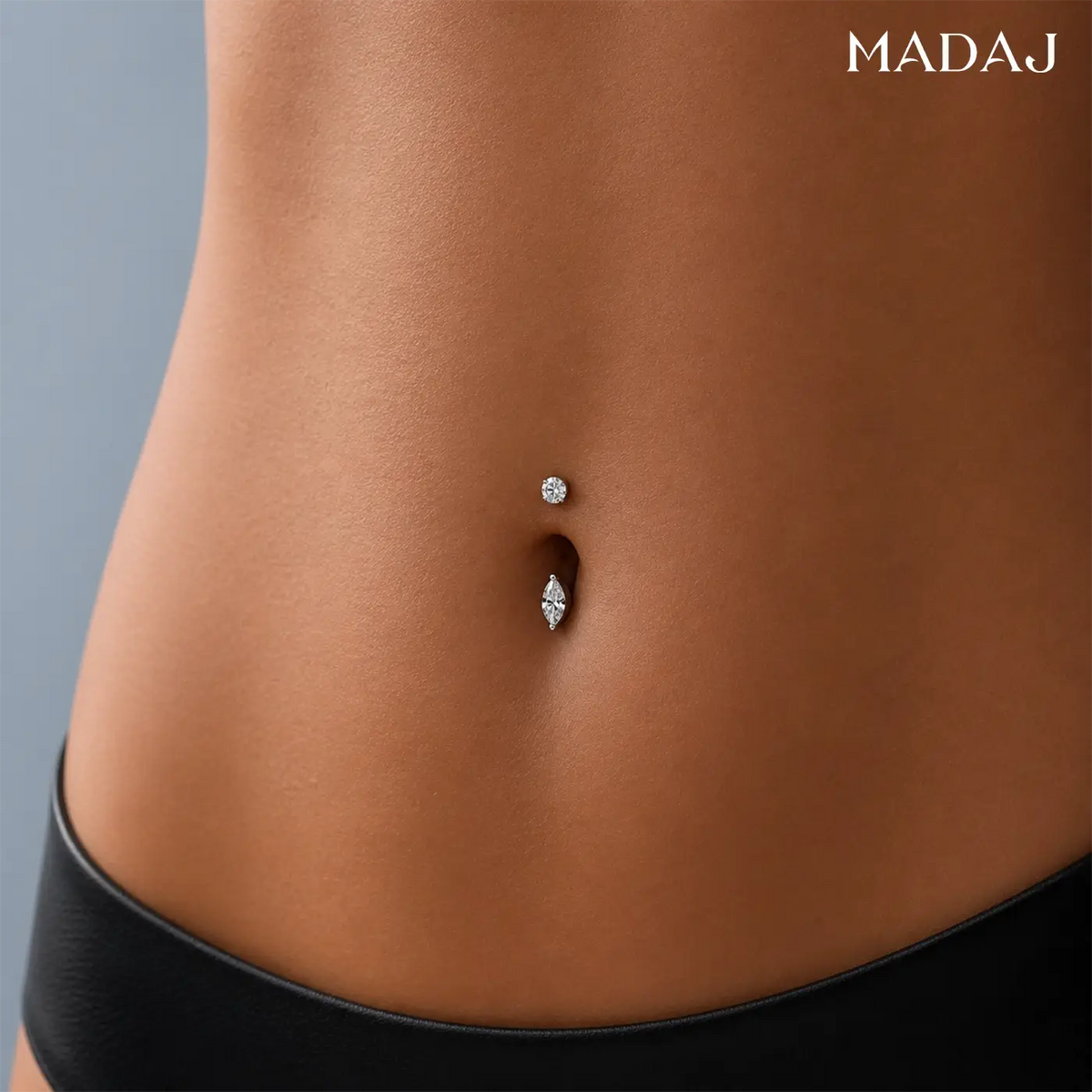 A model's midsection featuring a double navel piercing in white gold with a round diamond top and a marquise diamond bottom against a soft grey backdrop with the MADAJ logo in the corner.
