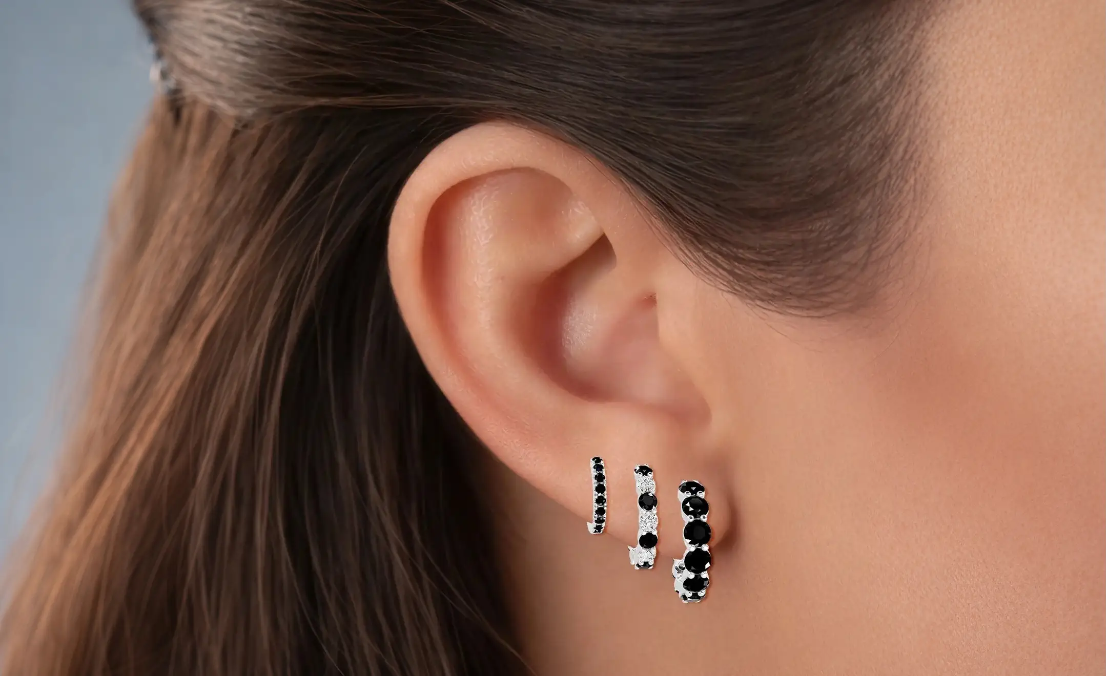 Three graduated size black diamond hoop earrings are shown worn on a person's ear, with the model's brown hair and ear visible against a soft gray background.