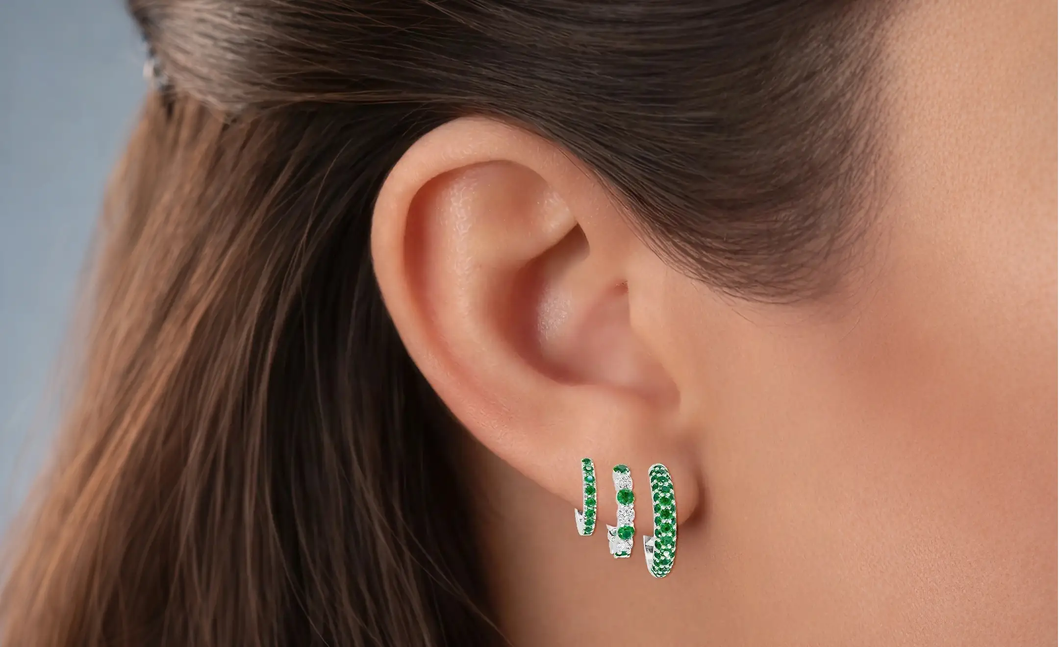 A close-up of a person's ear wearing three 18k white gold emerald hoop with diamond accent on the middle hoop worn on the lobe 