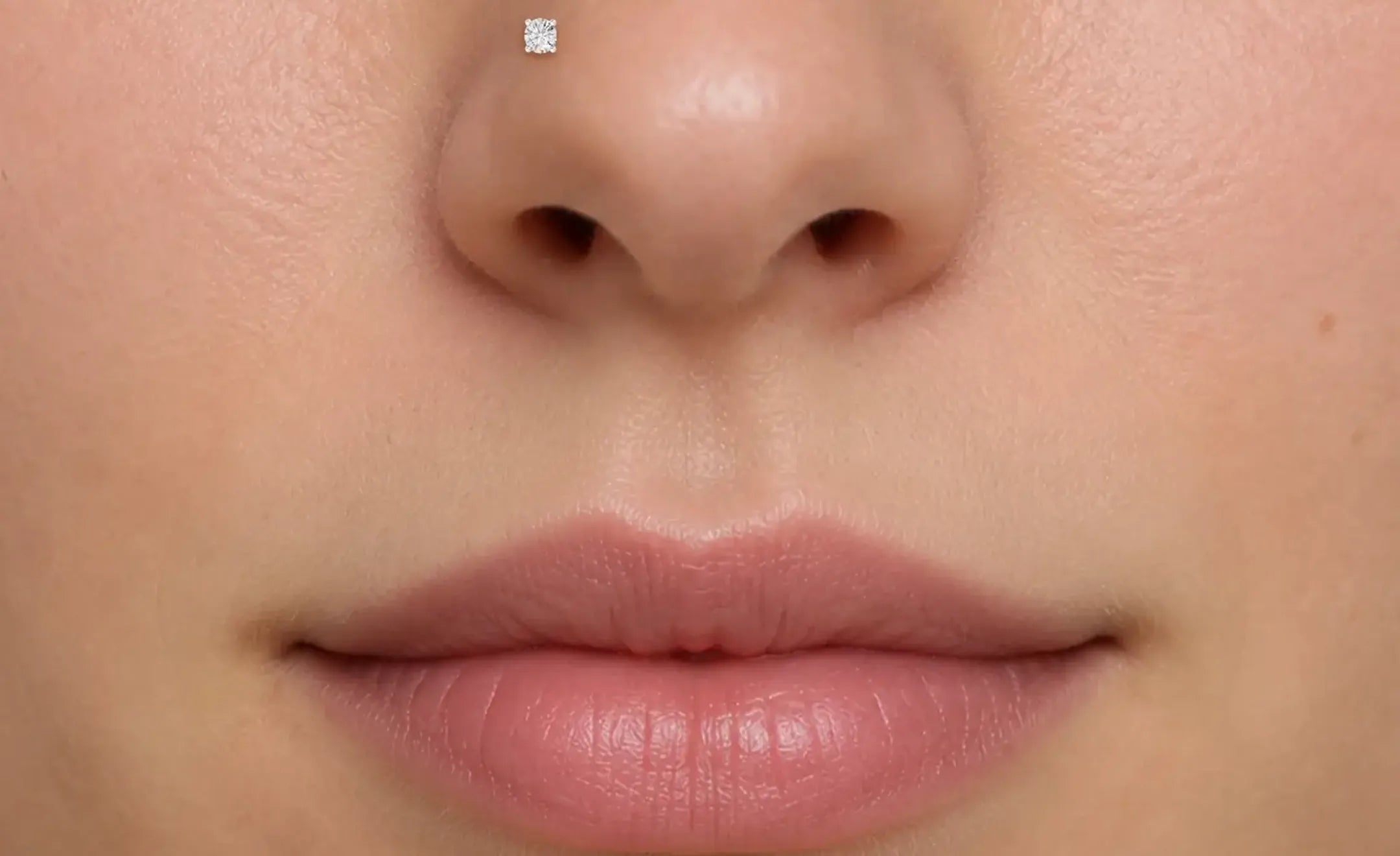 Extreme close-up of a person's nose and lips, showing a round diamond  stud nose piercing on the left side of the nose.
