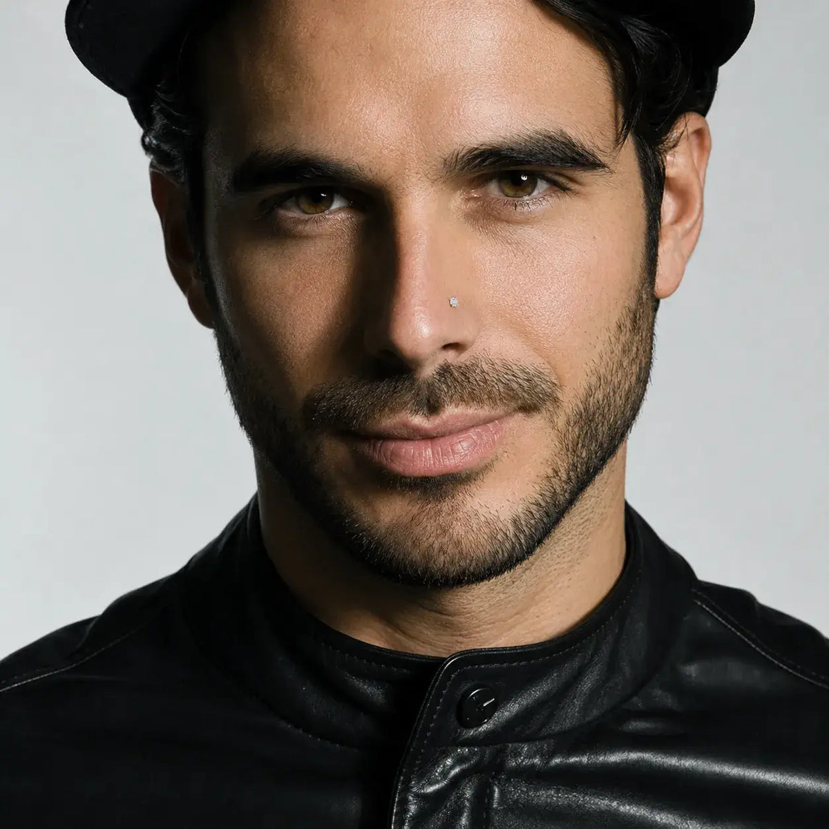Portrait of a male model in a black cap and leather jacket with a diamond nostril stud against a grey background.