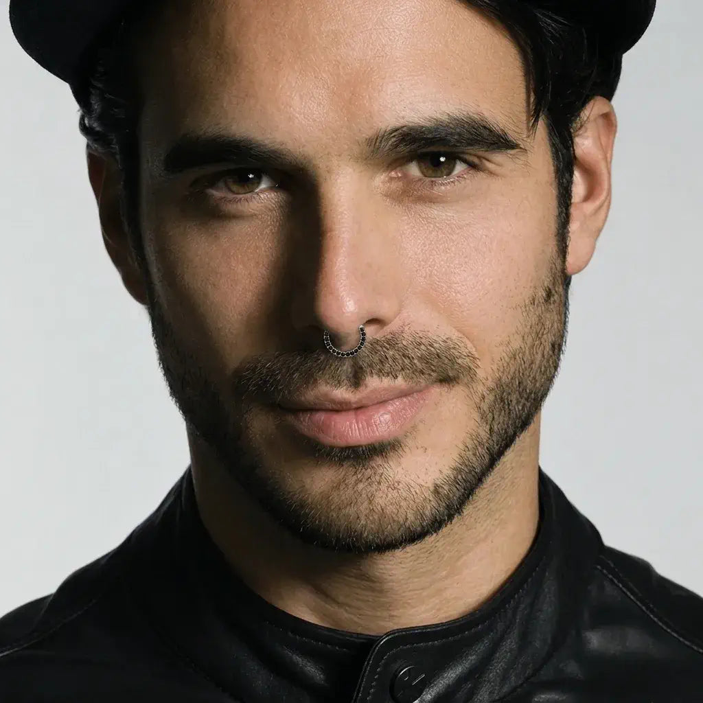 A close-up portrait of a man wearing a white gold black diamond septum ring, a black cap, and a black leather jacket against a light gray background.